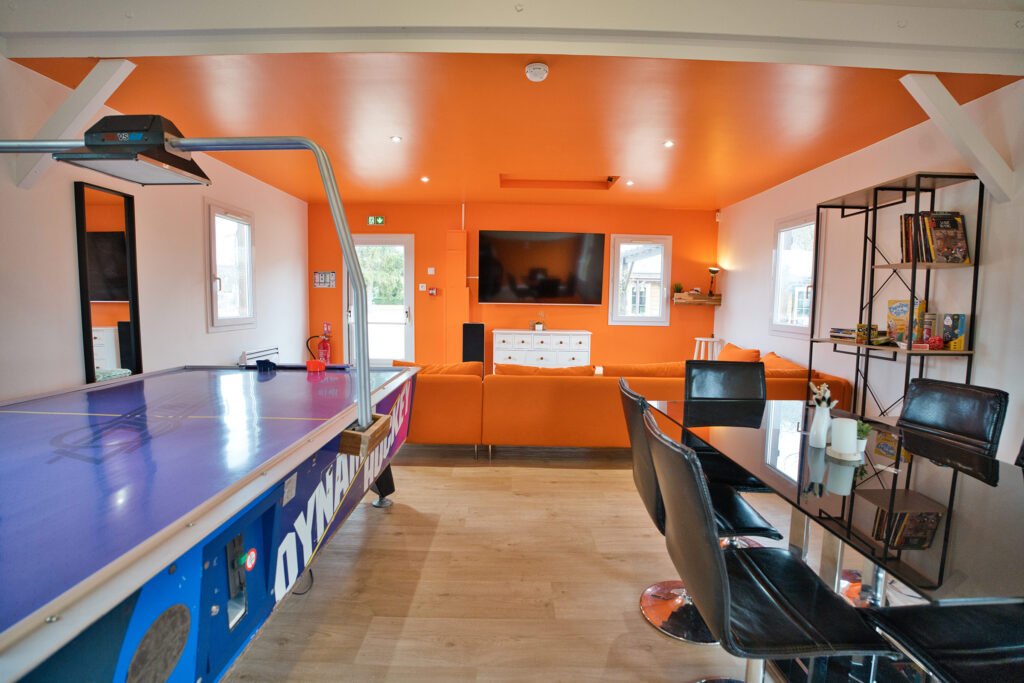 Salle de jeux du gîte Ranch de Sologne avec table de air hockey, table, canapé orange et télévision.
