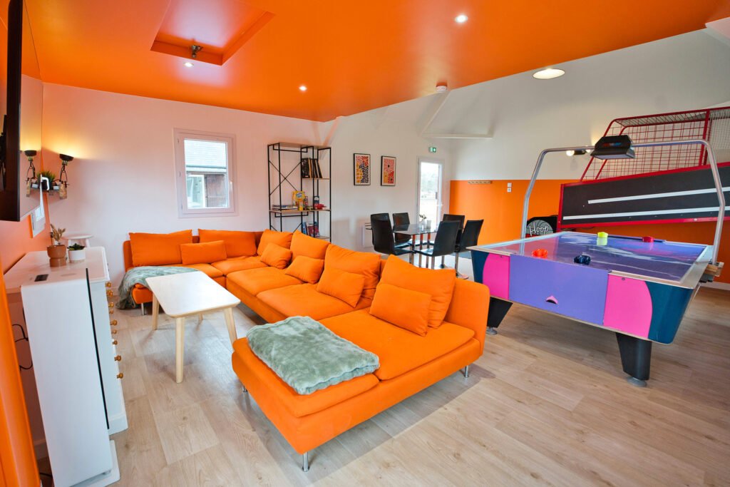 Salle de jeux du gîte Ranch de Sologne avec grand canapé orange, table de air hockey et espace détente.